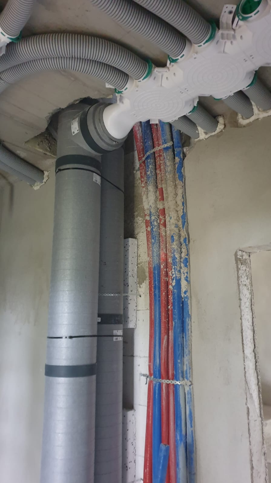 Montaż instalacji rekuperacji w surowym wnętrzu: szare rury wentylacyjne, biała rozdzielnica i czerwono-niebieskie rury, wszystko przymocowane do sufitu i ściany.