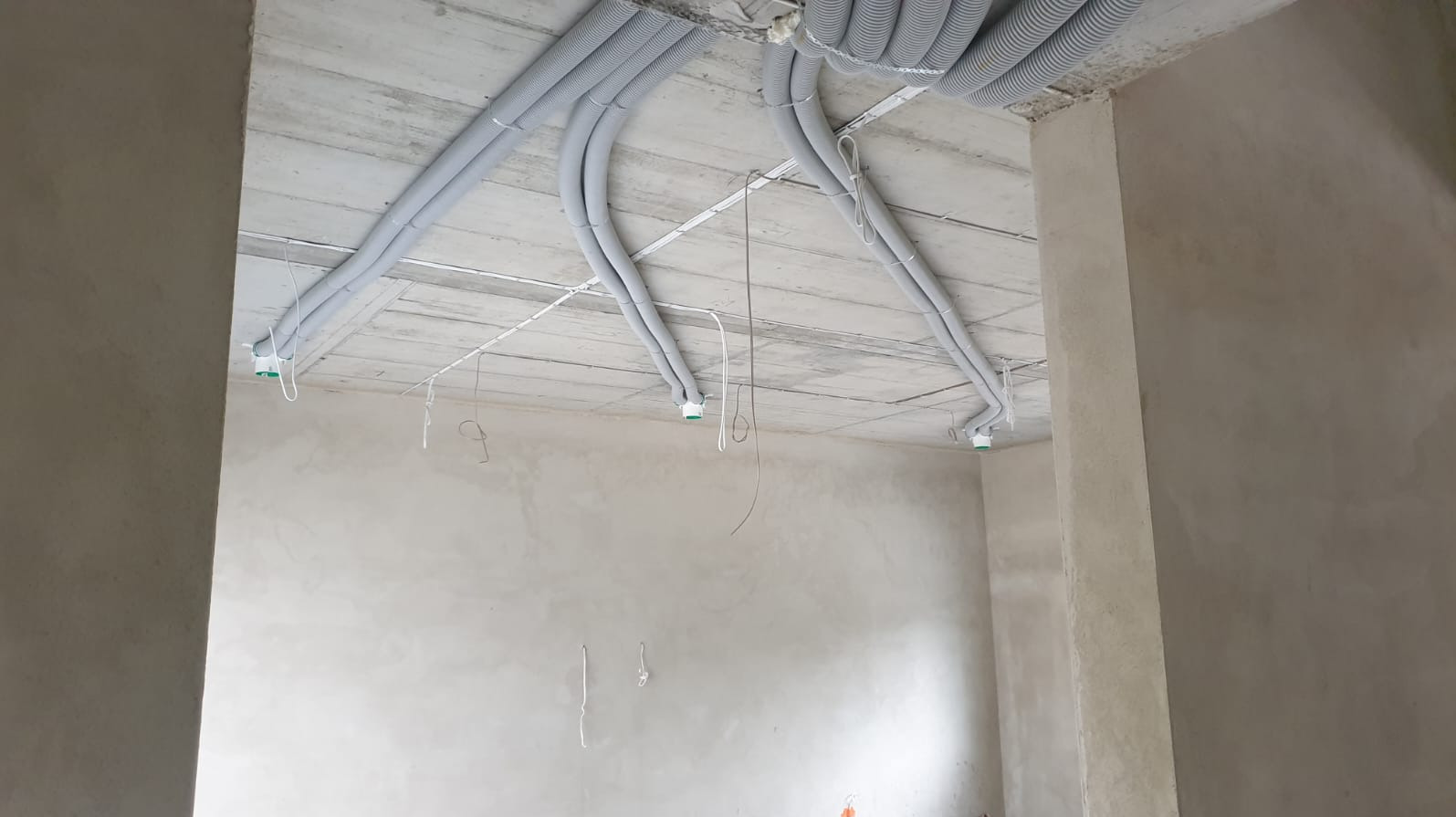 Szare, karbowane rury wentylacyjne pod betonowym sufitem w surowym wnętrzu. Widoczne przewody i uchwyty. Etap instalacji rekuperacji w nowym budynku.