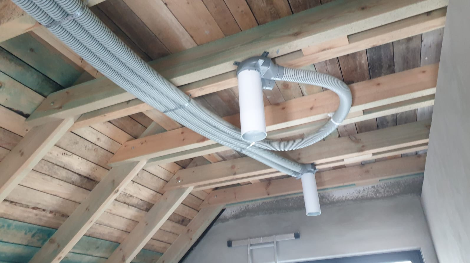Montaż systemu rekuperacji pod drewnianym stropem poddasza. Widoczne szare rury karbowane i białe elementy systemu wentylacyjnego.
