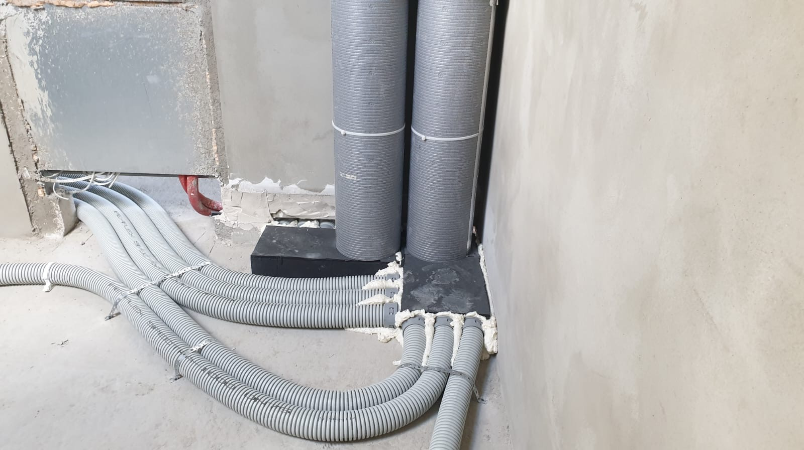 Szare, elastyczne rury wentylacyjne ułożone w podłodze i ścianie, uszczelnione pianką montażową, z widocznymi przewodami elektrycznymi i fragmentem czerwonej rury.