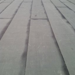 Usługi Dekarskie Maciej Włodarczyk - Szary, płaski dach pokryty papą bitumiczną w trakcie prac. Widoczne łączenia i fragmenty rolek papy. W tle osoba wykonująca prace dekarskie.