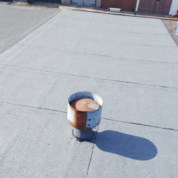 Usługi Dekarskie Maciej Włodarczyk - Szary, płaski dach pokryty papą z kominkiem wentylacyjnym na tle garaży. Widok z góry, oświetlenie dzienne.