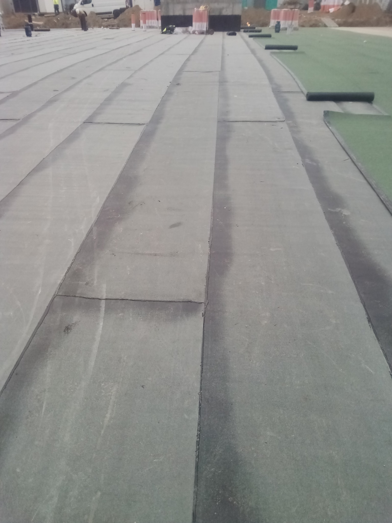 Szary dach pokryty papą termozgrzewalną, widoczne łączenia. Część dachu pokryta zieloną rolką. W tle ekipa budowlana i materiały budowlane. Ujęcie z góry.