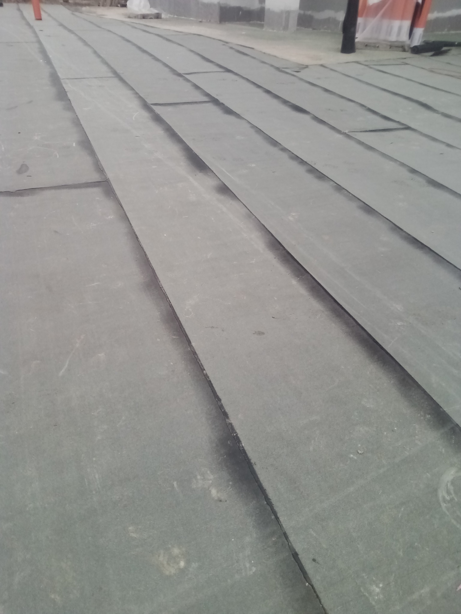 Szary, płaski dach pokryty papą bitumiczną, widok z góry. Widoczne łączenia papy i fragmenty konstrukcji dachu w tle. Dach po renowacji lub w trakcie prac.