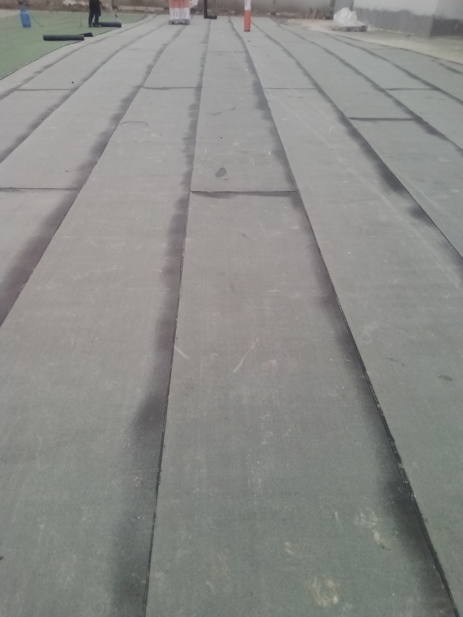 Szary, płaski dach pokryty papą bitumiczną w trakcie prac. Widoczne łączenia i fragmenty rolek papy. W tle osoba wykonująca prace dekarskie.