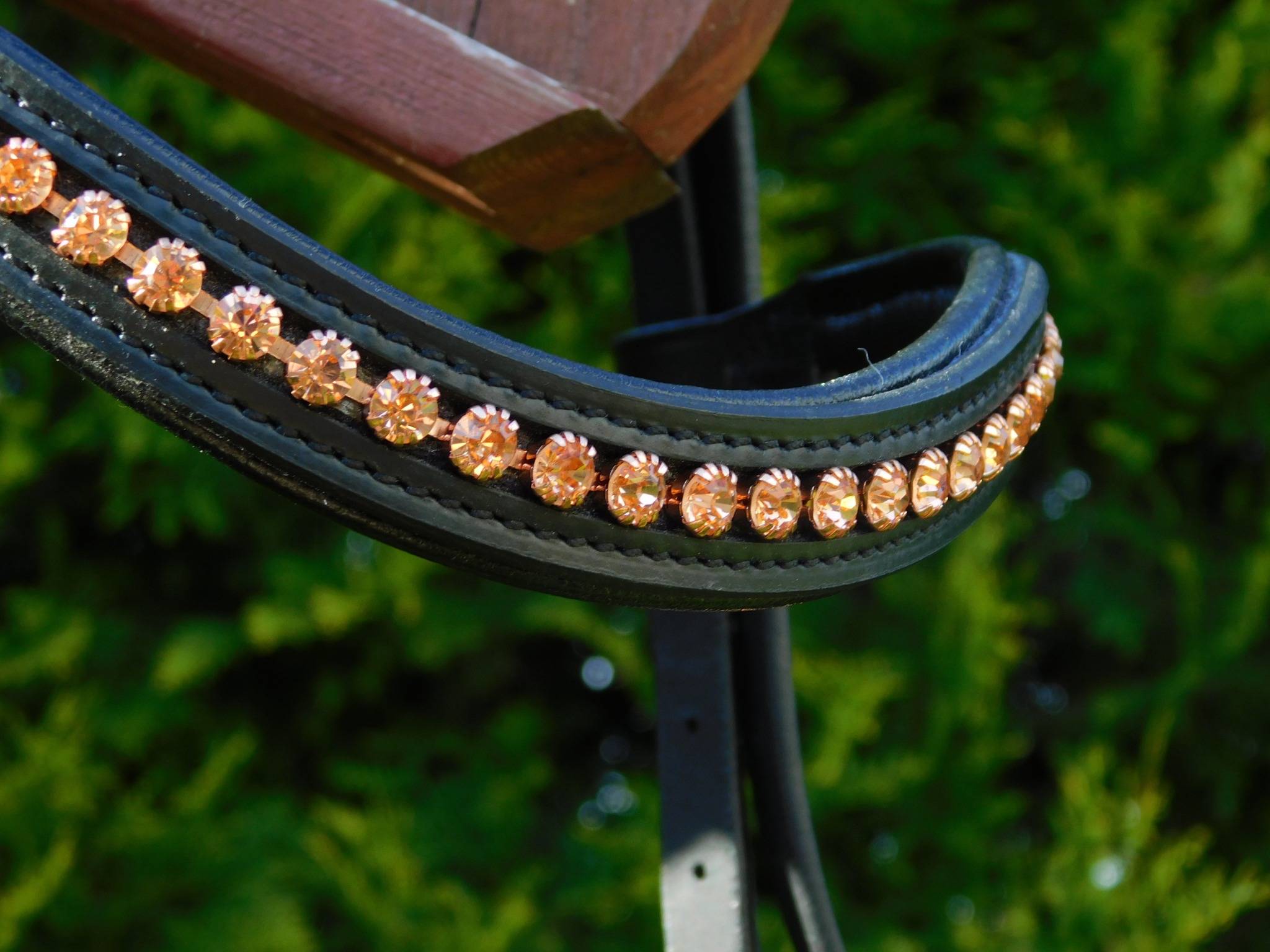 Ozdobne, czarne skórzane nagłowie końskie z rzędem błyszczących, pomarańczowych kryształków, na tle zieleni. Detal sprzętu jeździeckiego.