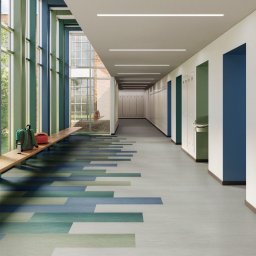 ZALENA GROUP SPÓŁKA Z OGRANICZONĄ ODPOWIEDZIALNOŚCIĄ - Korytarz szkolny z wykładziną w odcieniach błękitu i zieleni ułożoną w geometryczny wzór. Widoczne szafki i okno z plecakiem.