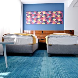 ZALENA GROUP SPÓŁKA Z OGRANICZONĄ ODPOWIEDZIALNOŚCIĄ - Błękitna wykładzina dywanowa w pokoju hotelowym z dwoma łóżkami i stolikiem. Geometryczny obraz na ścianie dodaje koloru.