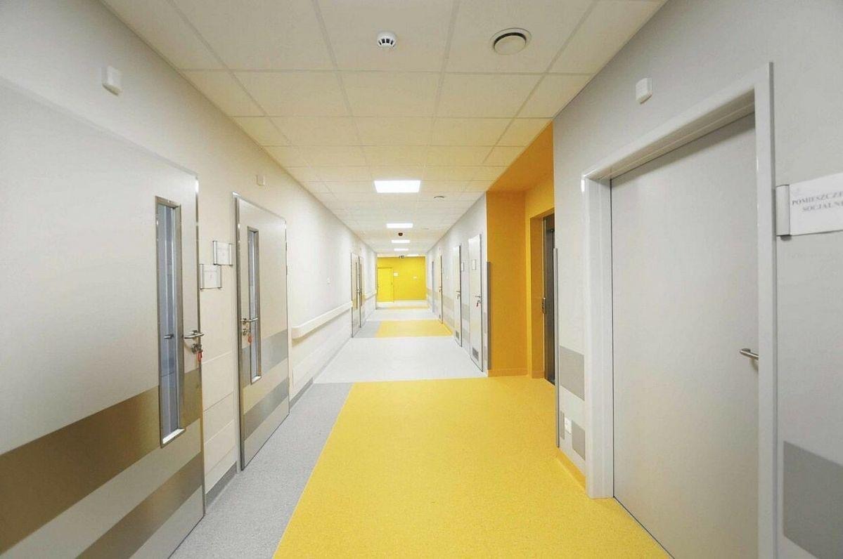 Długi korytarz w budynku użyteczności publicznej z żółtą wykładziną podłogową i białymi ścianami, oświetlony lampami sufitowymi. Widoczne drzwi i listwy przypodłogowe.