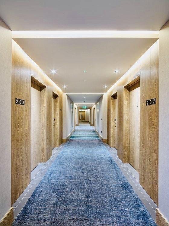 Długi korytarz hotelowy z niebieską wykładziną dywanową. Drzwi pokojowe z numerami 207 i 208 po bokach. Oświetlenie sufitowe nadaje ciepły blask.