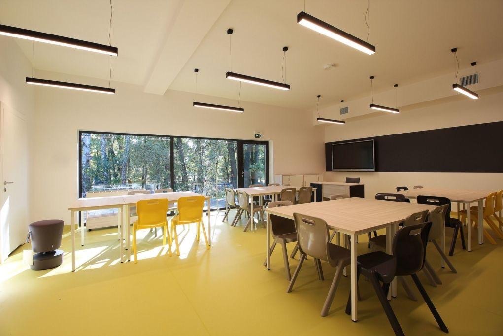 Żółta podłoga w jasnej sali z oknami. Stoły i krzesła ustawione w rzędach, nowoczesne oświetlenie sufitowe. Widok na zieleń za oknami.