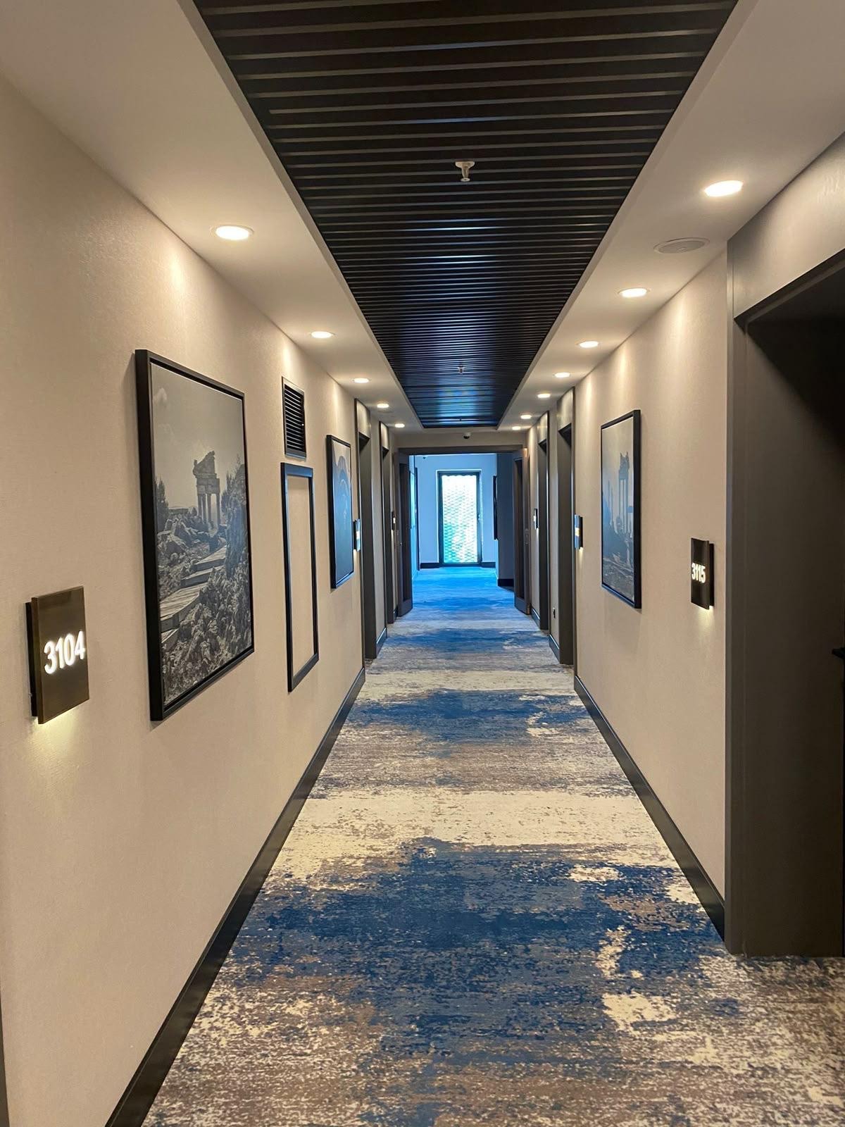 Długi korytarz hotelowy z wykładziną w abstrakcyjny wzór w odcieniach błękitu i beżu. Na ścianach obrazy w czarnych ramach i numery pokoi z podświetleniem.