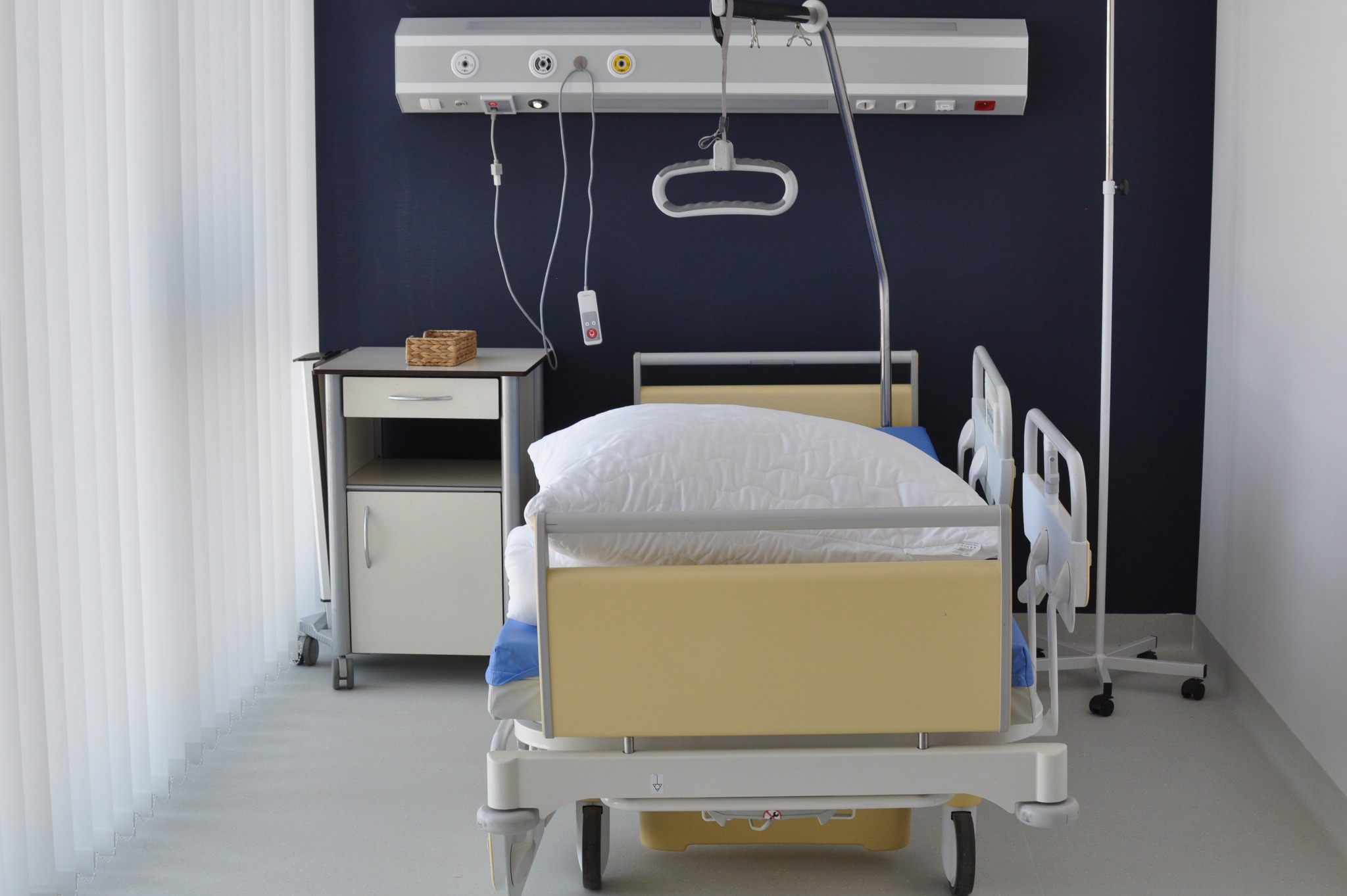 Jasna, pusta sala szpitalna z łóżkiem na kółkach, szafką nocną i aparaturą medyczną na ścianie. Widoczne pionowe żaluzje i ciemna ściana w tle.