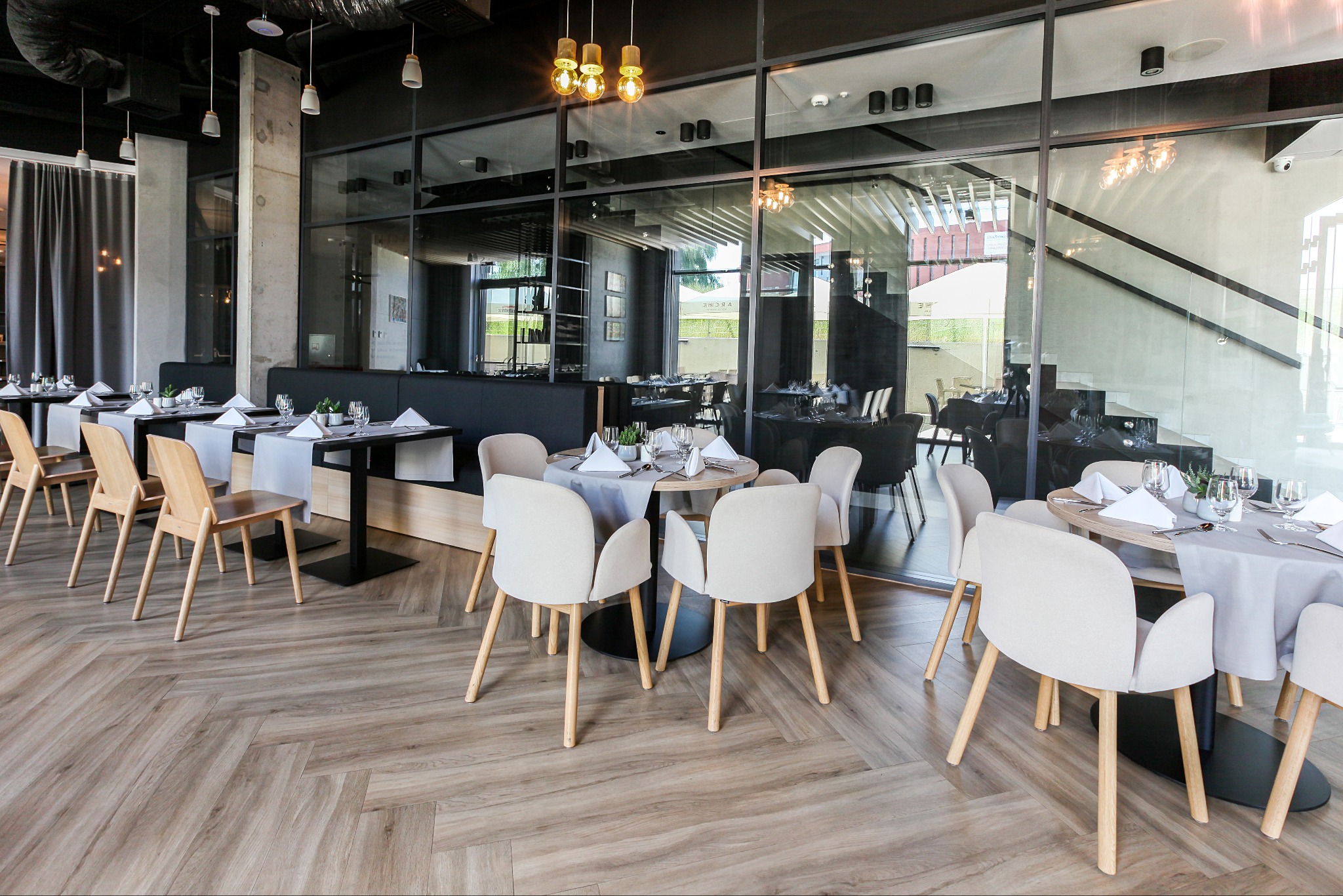 Jasna, drewnopodobna podłoga ułożona w jodełkę francuską w eleganckiej restauracji. Widoczne stoły z nakryciami i krzesła w neutralnych barwach.