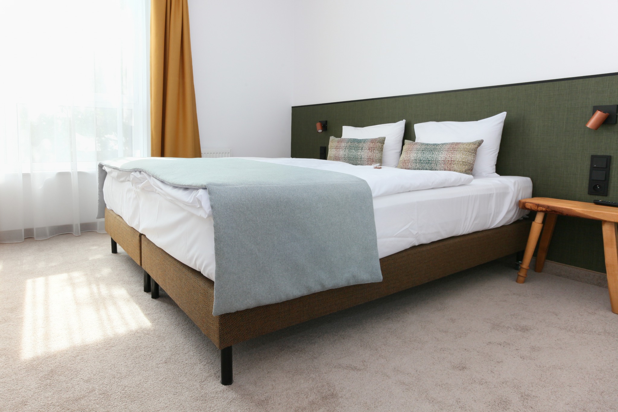 Jasny pokój hotelowy z łóżkiem, wykładziną dywanową i drewnianym stolikiem nocnym. Widok na okno z zasłonami. Neutralna kolorystyka i miękkie światło.