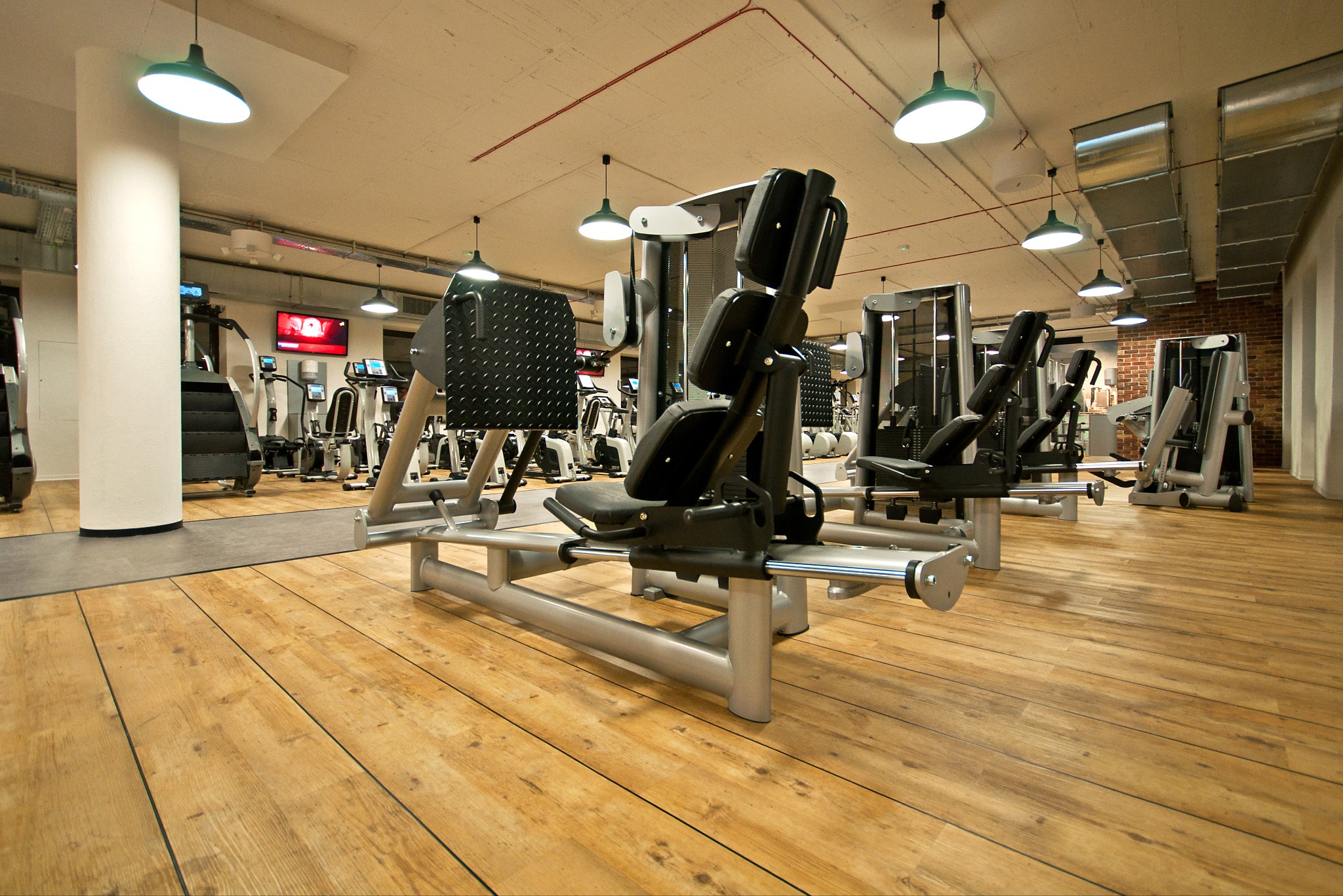 Jasna, drewniana podłoga w siłowni z maszynami do ćwiczeń, oświetlona wiszącymi lampami. Widok z niskiej perspektywy podkreśla głębię przestrzeni.