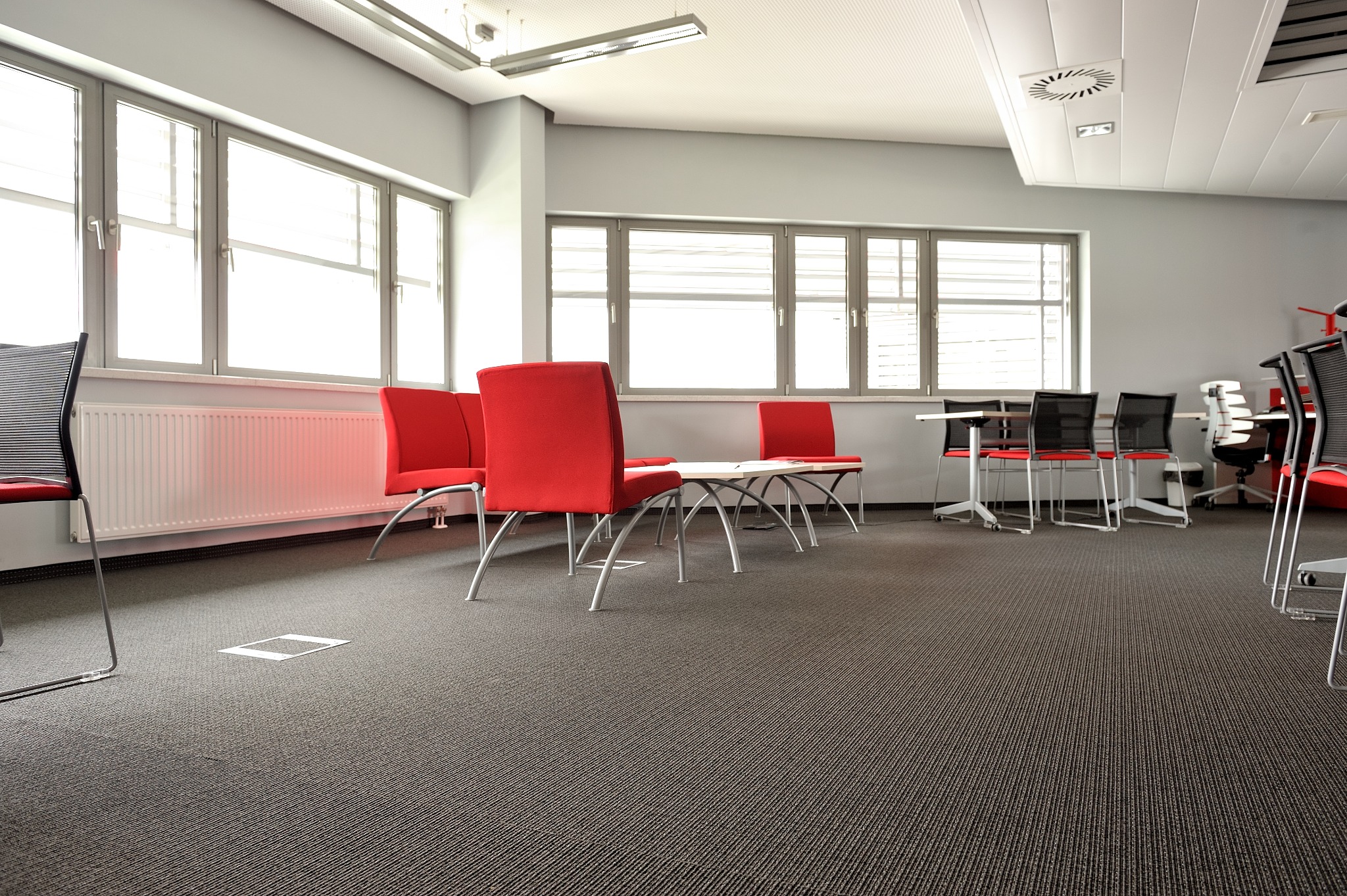 Biuro z szarą wykładziną dywanową w jodełkę, czerwonymi krzesłami i dużymi oknami. Widoczny fragment podłogi z dostępem do instalacji.