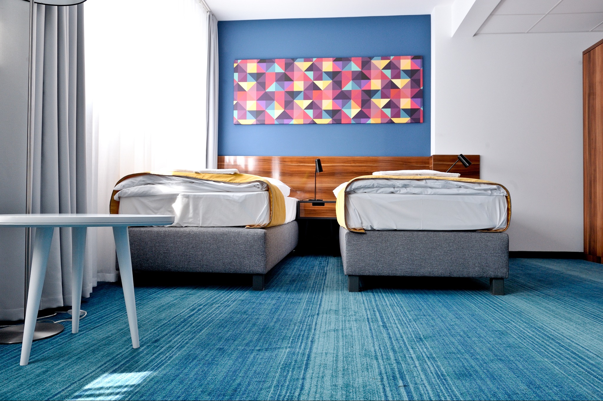 Błękitna wykładzina dywanowa w pokoju hotelowym z dwoma łóżkami i stolikiem. Geometryczny obraz na ścianie dodaje koloru.