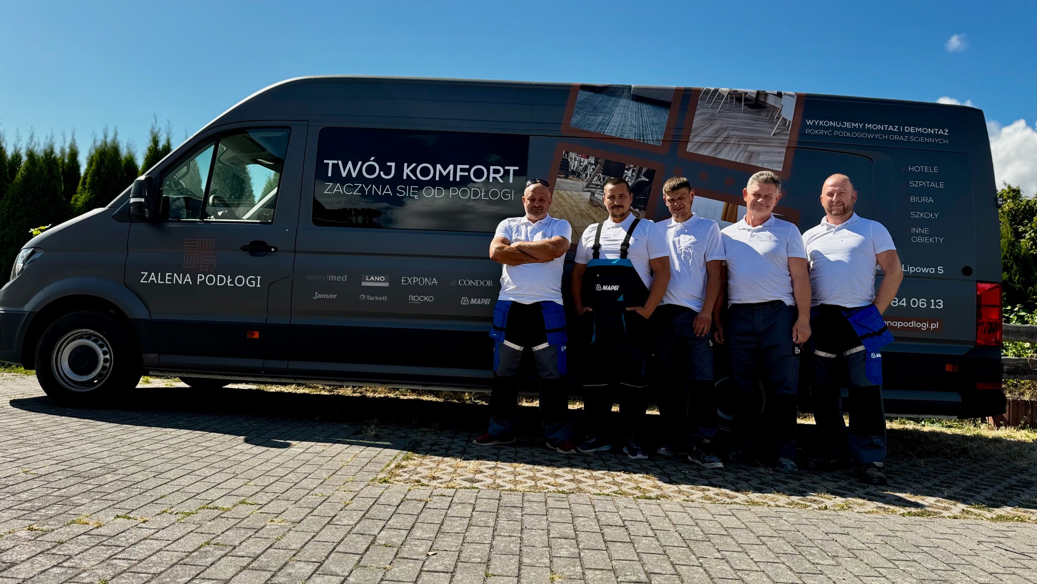 Ekipa monterska na tle firmowego busa z reklamą usług podłogowych: montaż, demontaż. Widoczne logo firmy i dane kontaktowe. Dzień słoneczny.