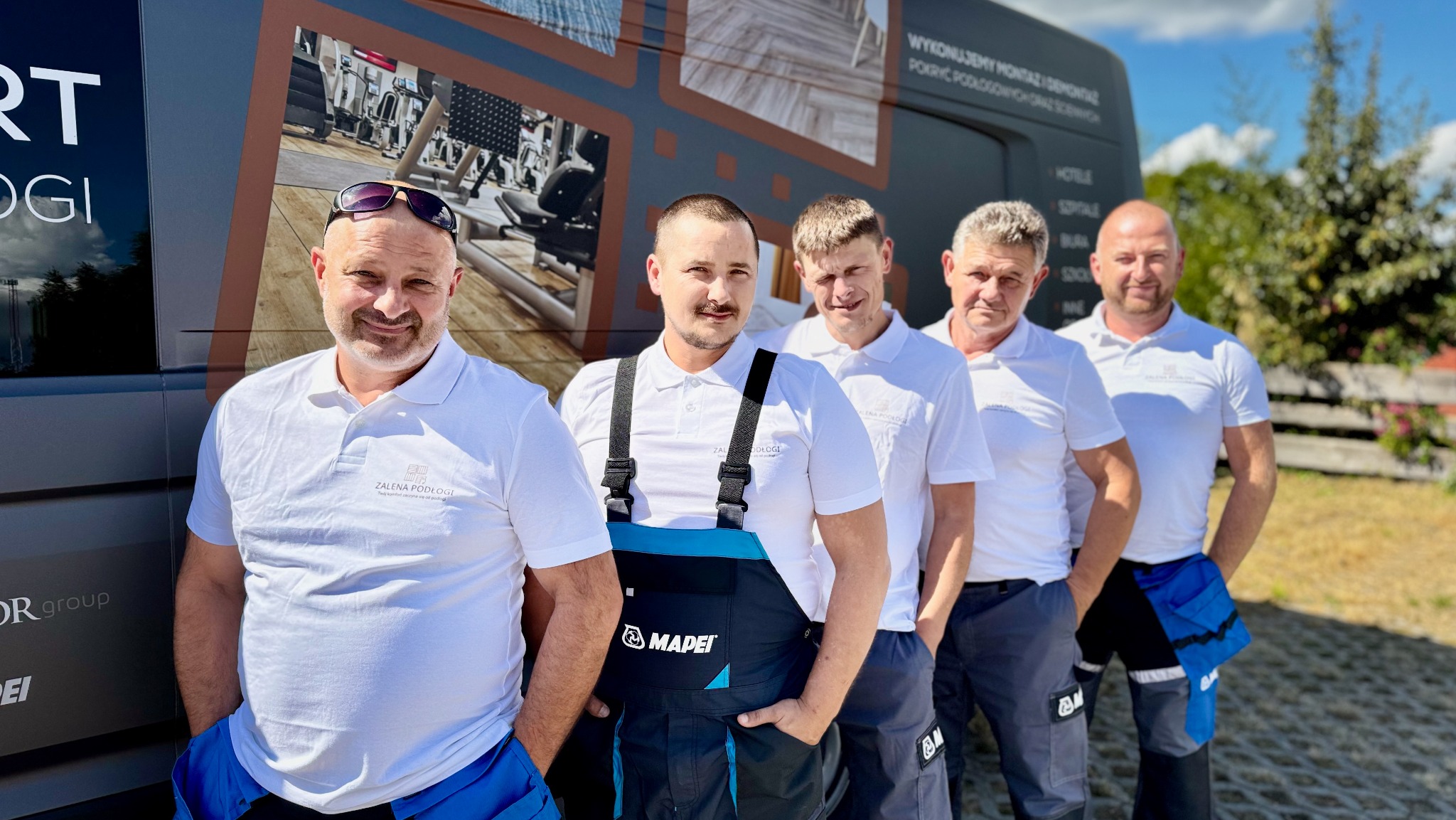 Zespół monterów podłóg 'Zalena Podłogi' pozuje przed firmowym busem z reklamą usług montażu w obiektach hotelowych i biurowych. Ubrani w firmowe polo i robocze spodnie.