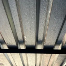 Blaszany dach z widocznymi kroplami wody, sfotografowany pod kątem od dołu. Widoczne elementy konstrukcji dachu i odbicia światła na powierzchni blachy.