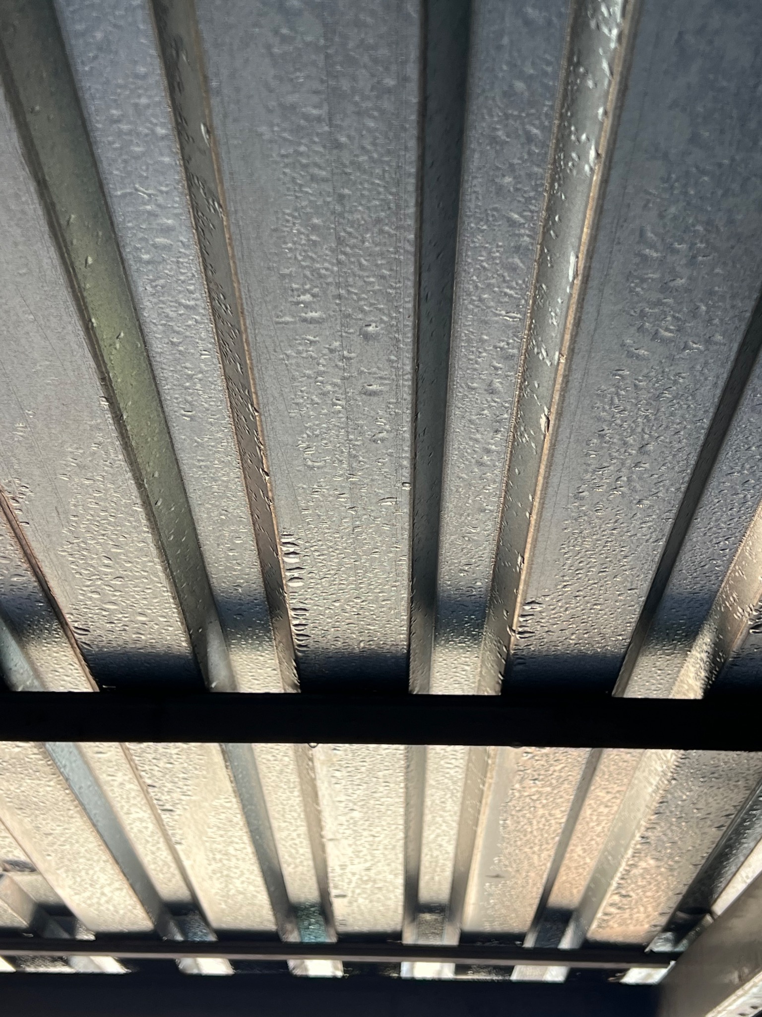 Blaszany dach z widocznymi kroplami wody, sfotografowany pod kątem od dołu. Widoczne elementy konstrukcji dachu i odbicia światła na powierzchni blachy.