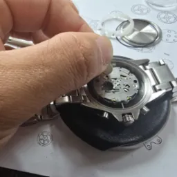 Wymiana baterii w zegarku na metalowej bransolecie. Widoczny mechanizm zegarka, narzędzia i schemat zegarka na papierze. Precyzyjna praca zegarmistrza.