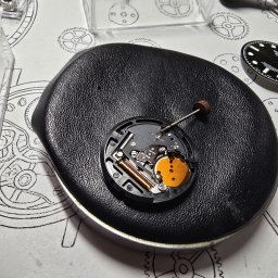 ABC MULTIMEDIA MICHAŁ KAPAŁKA - Otwarty mechanizm zegarka na czarnym podłożu, obok tarcza i narzędzia zegarmistrzowskie na papierze z rysunkami trybów. Praca zegarmistrza.