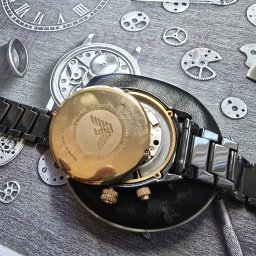 ABC MULTIMEDIA MICHAŁ KAPAŁKA - Zegarek Emporio Armani z otwartym deklem na czarnym podłożu z nadrukowanymi elementami zegarków. Widoczne narzędzia zegarmistrzowskie, detale mechanizmu i złote elementy.