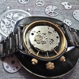 ABC MULTIMEDIA MICHAŁ KAPAŁKA - Otwarty, metalowy zegarek na skórzanej podkładce z widocznym mechanizmem i baterią, na tle z grafiką zegarków i trybików. Precyzyjna robota zegarmistrzowska.