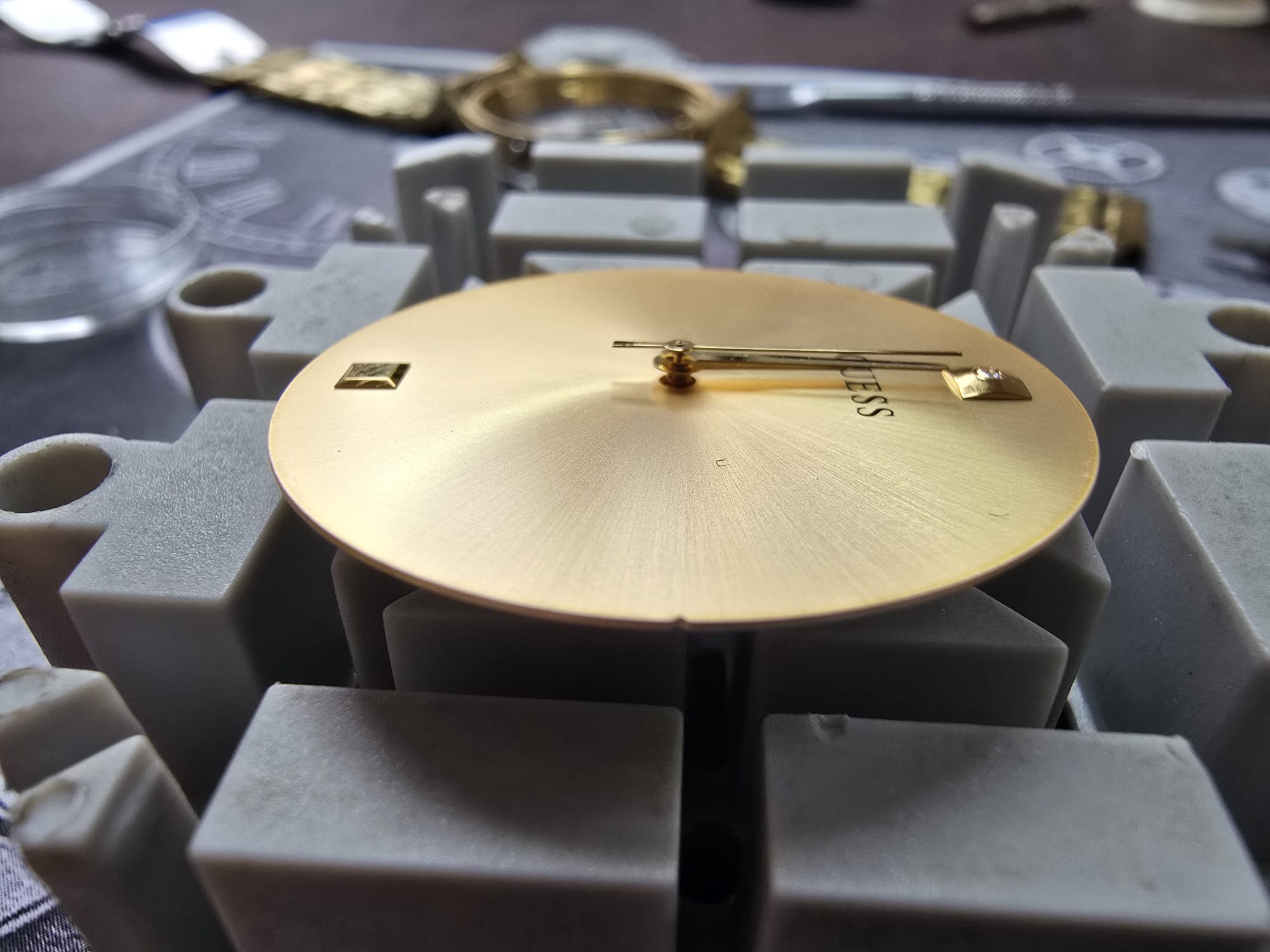 Złota tarcza zegarka Guess w trakcie naprawy, osadzona na szarym uchwycie. Widoczne narzędzia zegarmistrzowskie w tle, co sugeruje precyzyjną pracę serwisową.