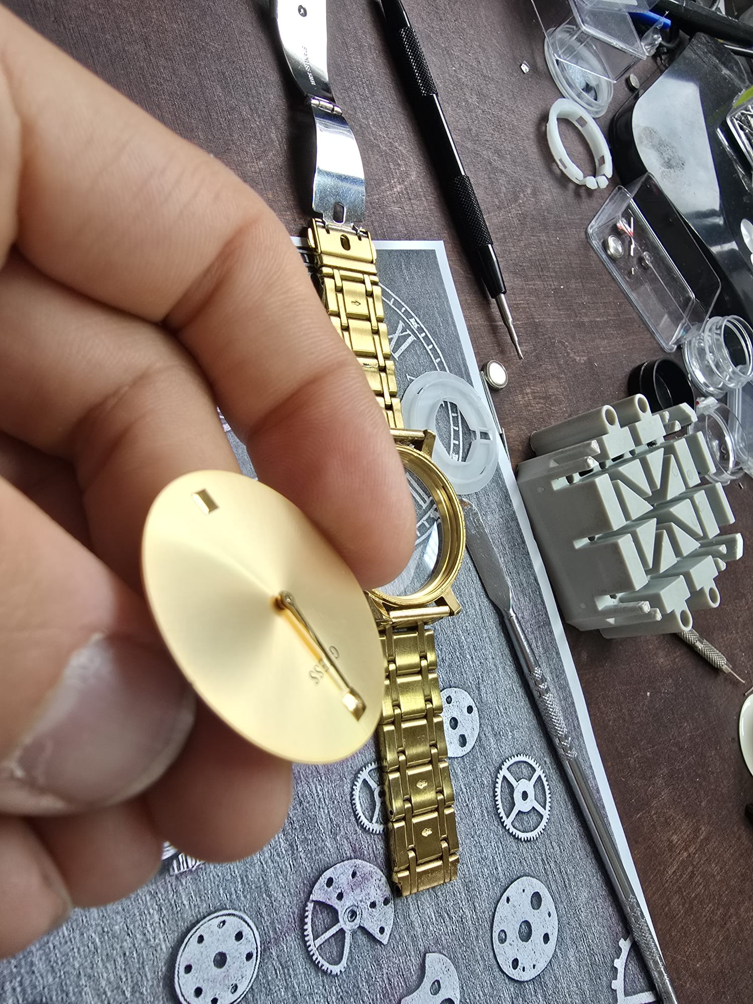 Złoty zegarek marki Guess w trakcie naprawy. Widoczna tarcza w dłoni, narzędzia zegarmistrzowskie i schemat mechanizmu na tle. Precyzyjna praca zegarmistrza.