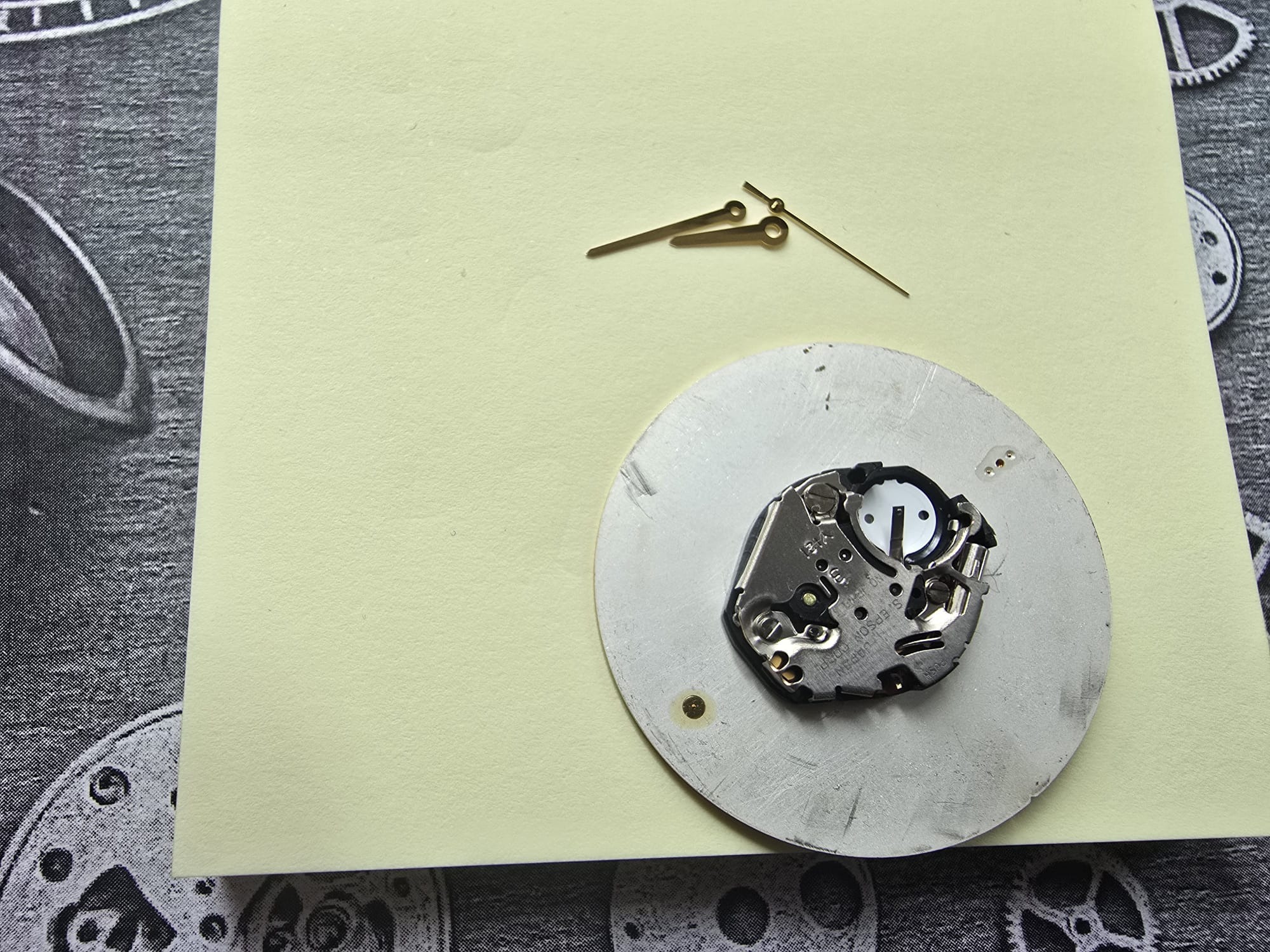 Mechanizm zegarka kwarcowego z trzema wskazówkami na żółtej kartce, na tle wzorzystej tkaniny. Widoczne detale mechanizmu.