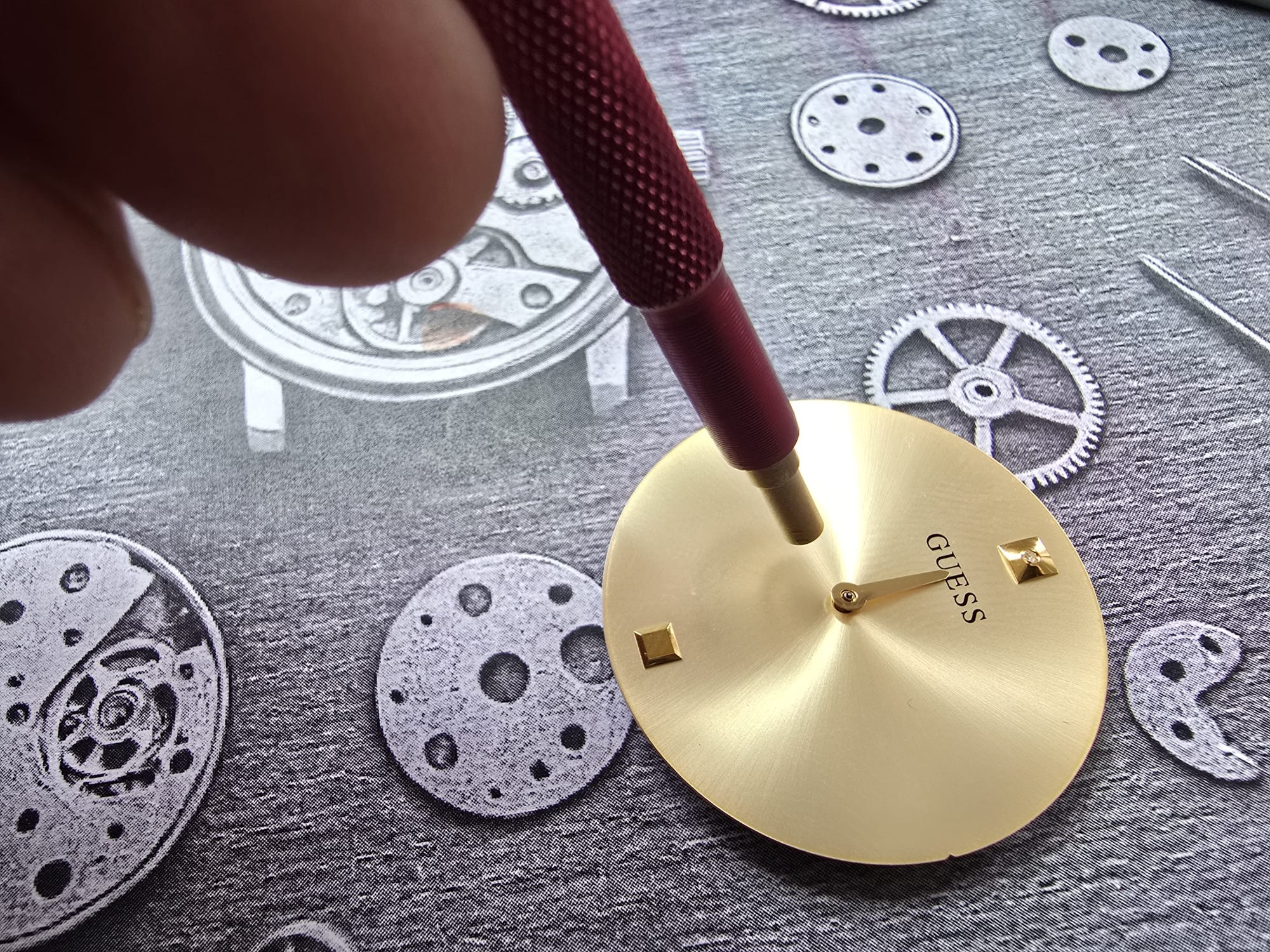 Naprawa zegarka: narzędzie precyzyjne nad złotą tarczą zegarka Guess, na tle z elementami mechanizmu i schematami. Detal naprawy zegarmistrzowskiej.