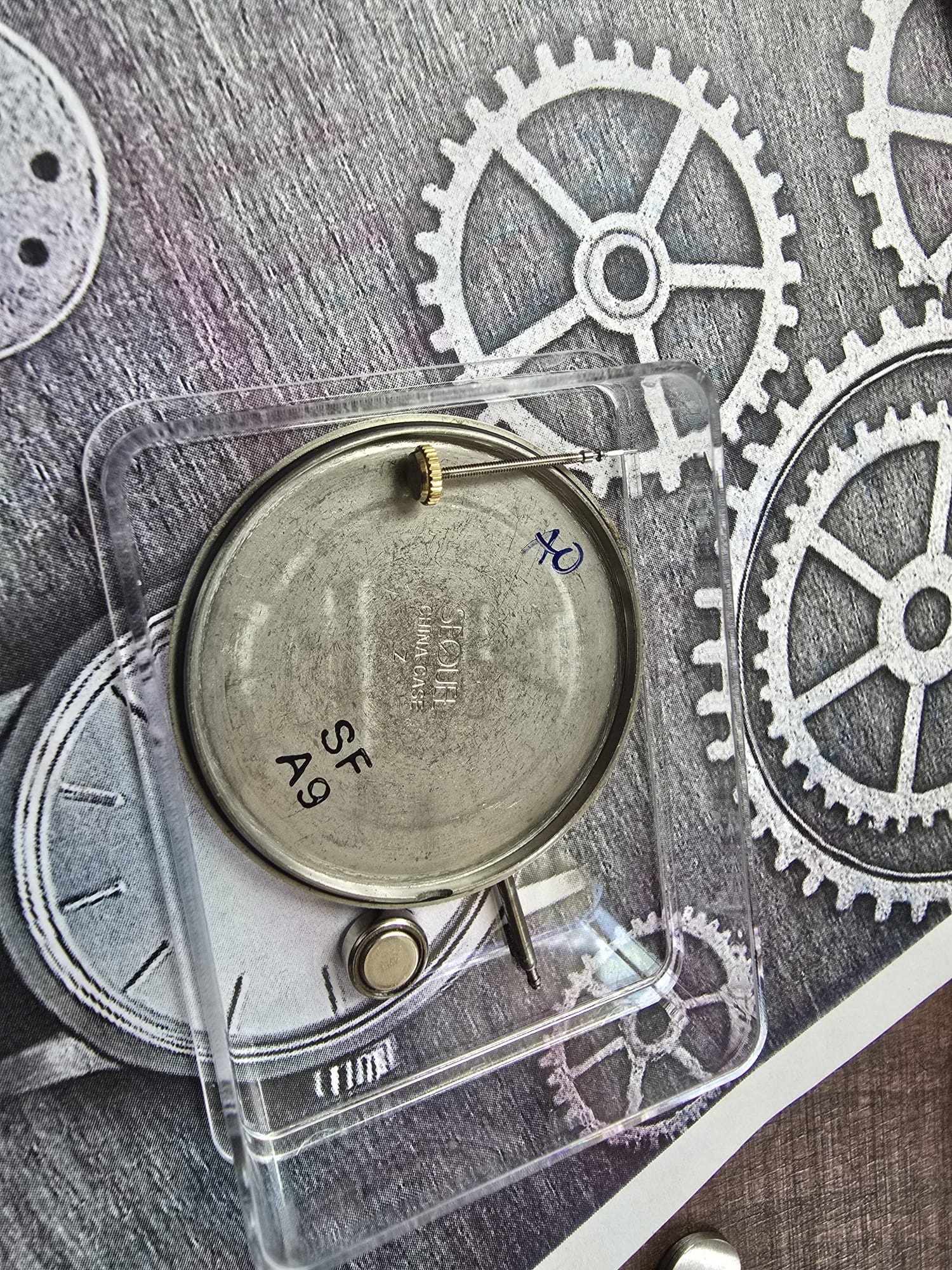 Otwarta obudowa zegarka w przezroczystym pudełku na tle grafiki z zębatkami, widoczny mechanizm i bateria. Napisy na obudowie zegarka.