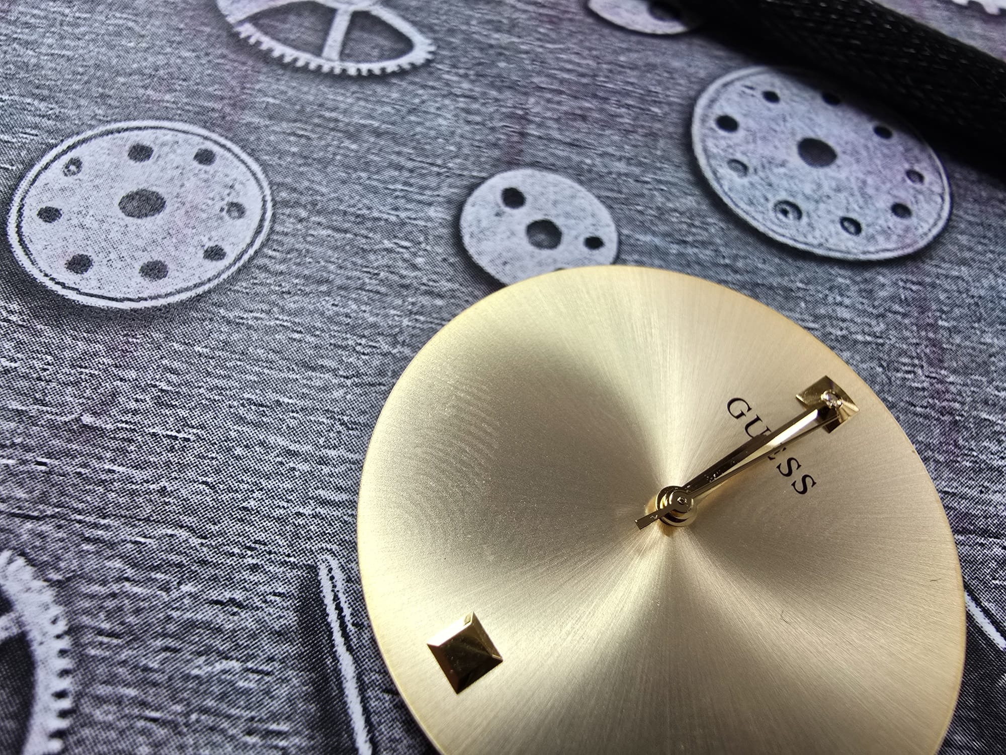 Złota tarcza zegarka Guess z wskazówkami na tle z motywem zębatek. Detal zegarmistrzowski w zbliżeniu, prezentujący precyzję i elegancję. Naprawa zegarków.