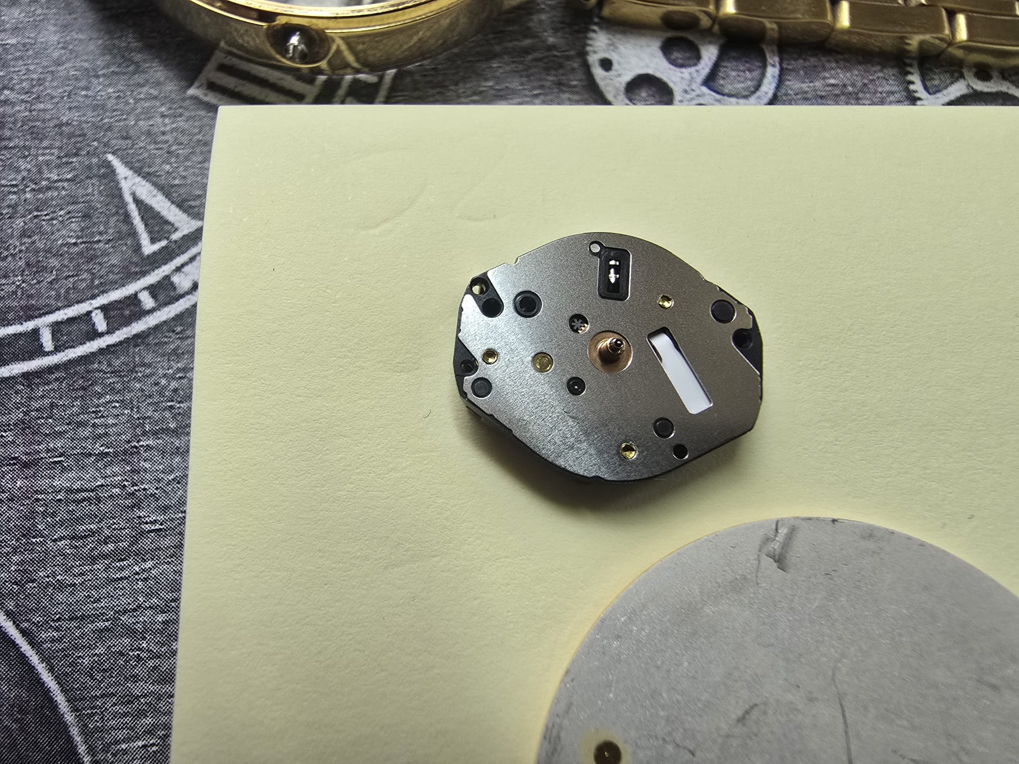 Precyzyjny mechanizm kwarcowy zegarka na beżowym tle, obok fragmenty zegarka i narzędzi. Detaliczna prezentacja komponentu podczas serwisu zegarmistrzowskiego.