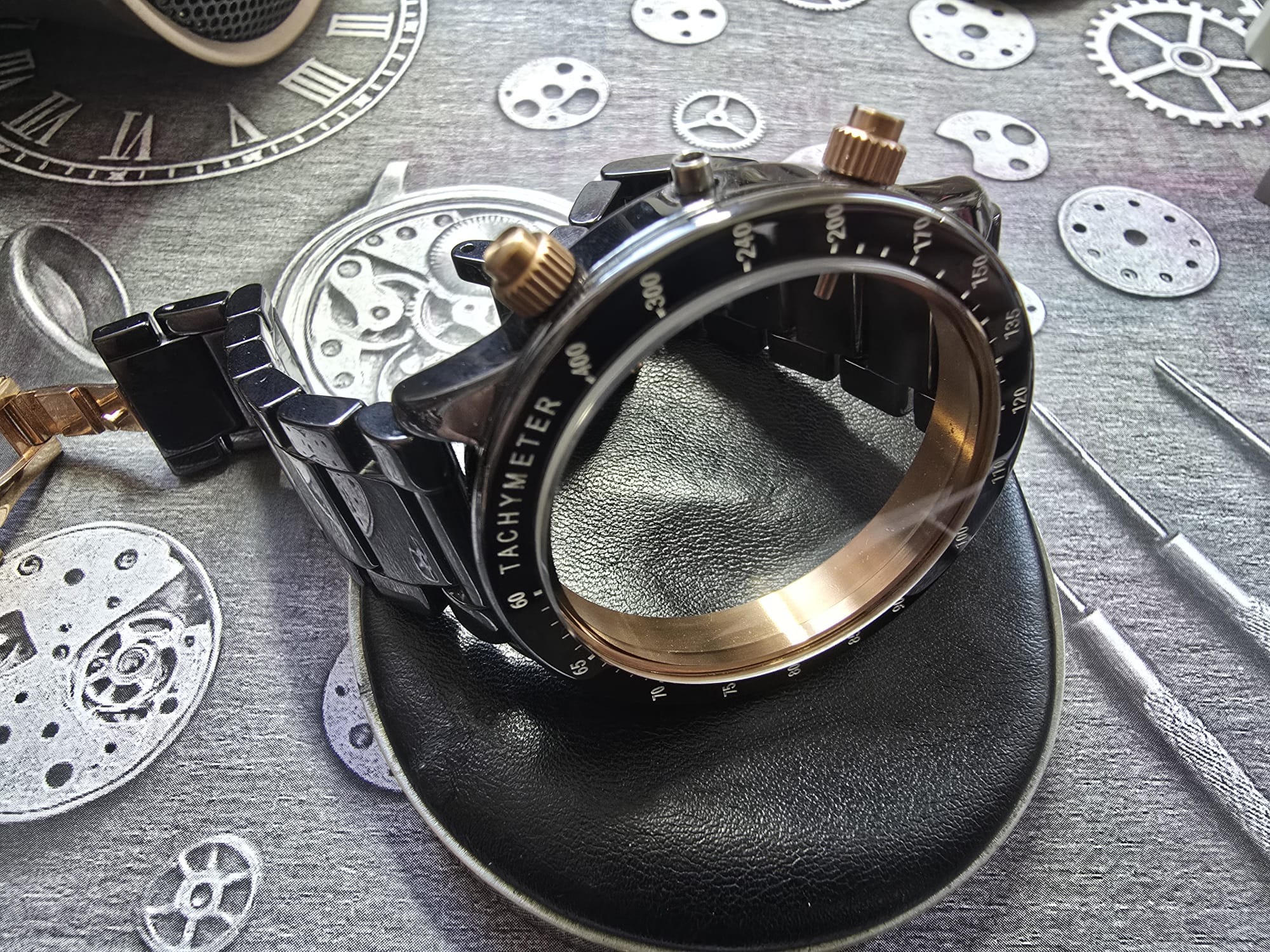 Czarny zegarek z tachymetrem, bez szkiełka, leżący na czarnym, skórzanym podłożu. W tle narzędzia i grafika z mechanizmami zegarków.