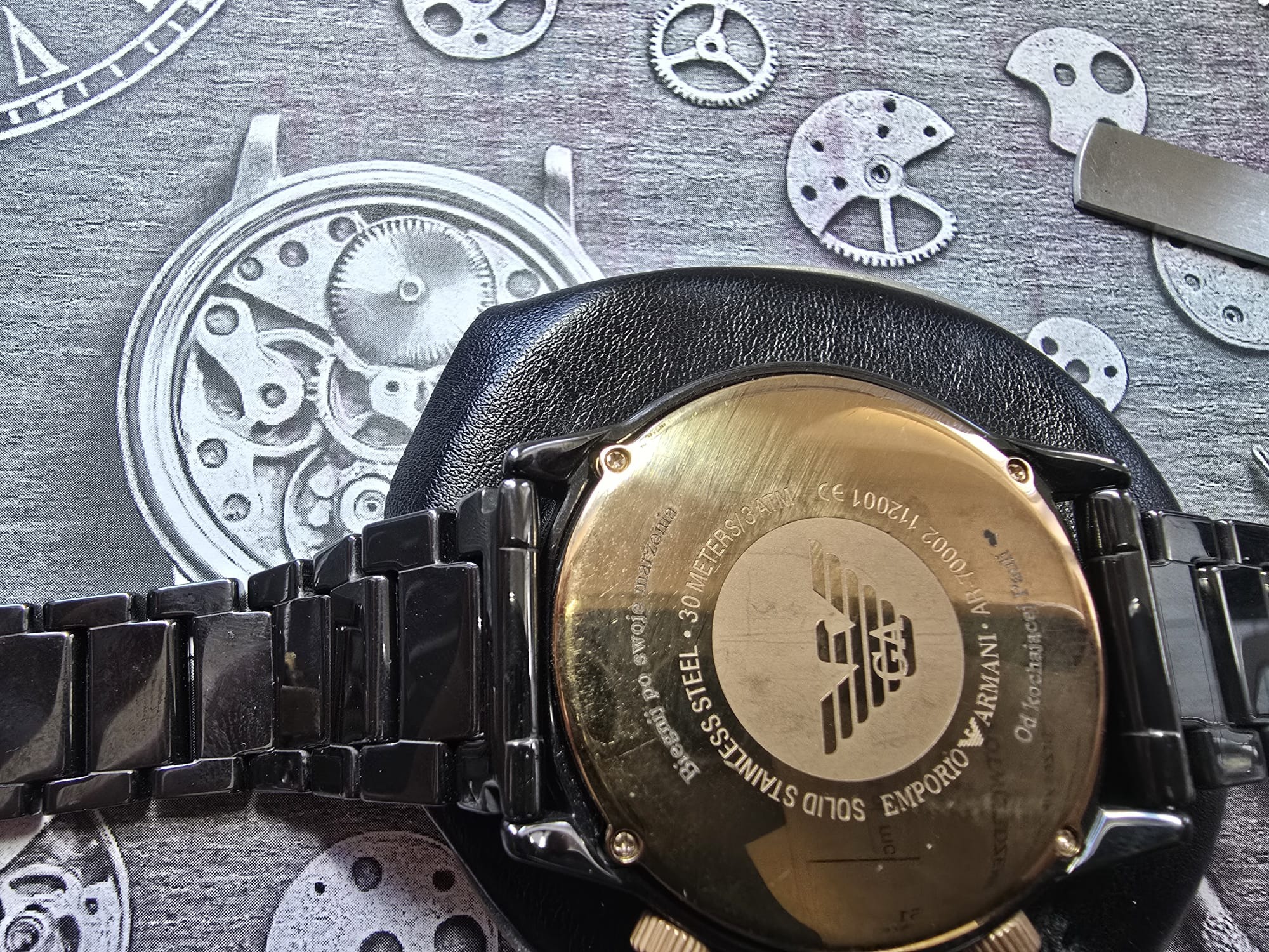Czarny zegarek Emporio Armani z widocznym deklem i elementami mechanizmu w tle. Metalowa bransoleta i złoty dekiel z logo marki. Detaliczny widok na precyzję wykonania.