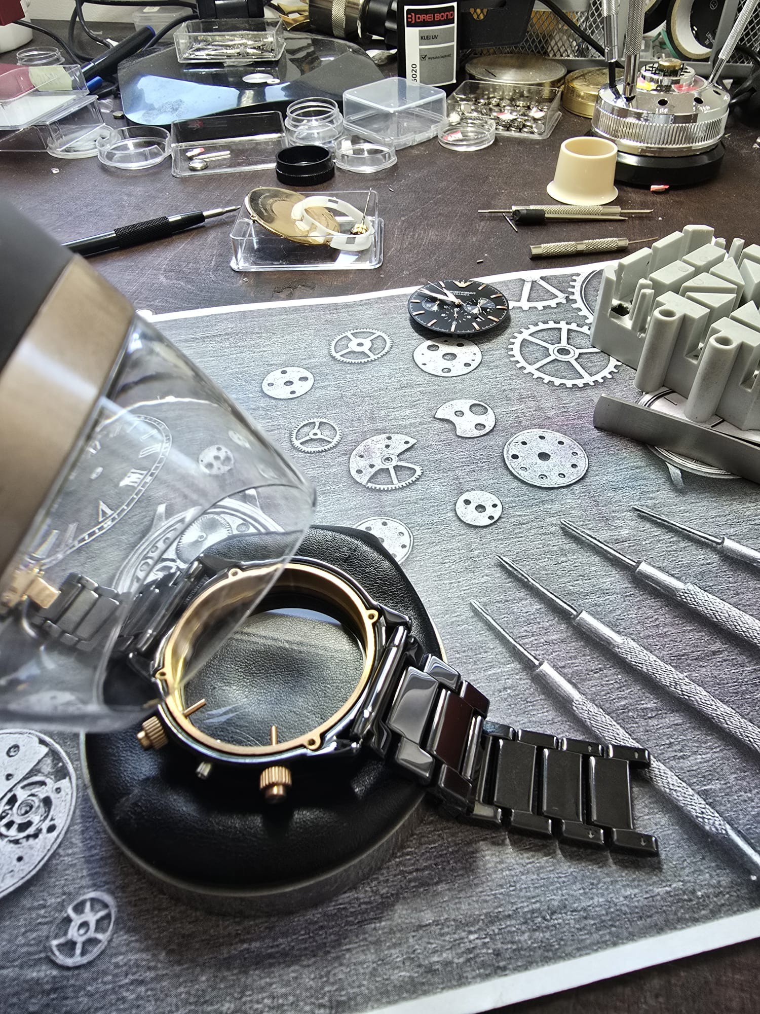 Zegarek z czarną bransoletą w trakcie naprawy, elementy mechanizmu i narzędzia na tle z grafiką trybów. Precyzyjna praca zegarmistrzowska.