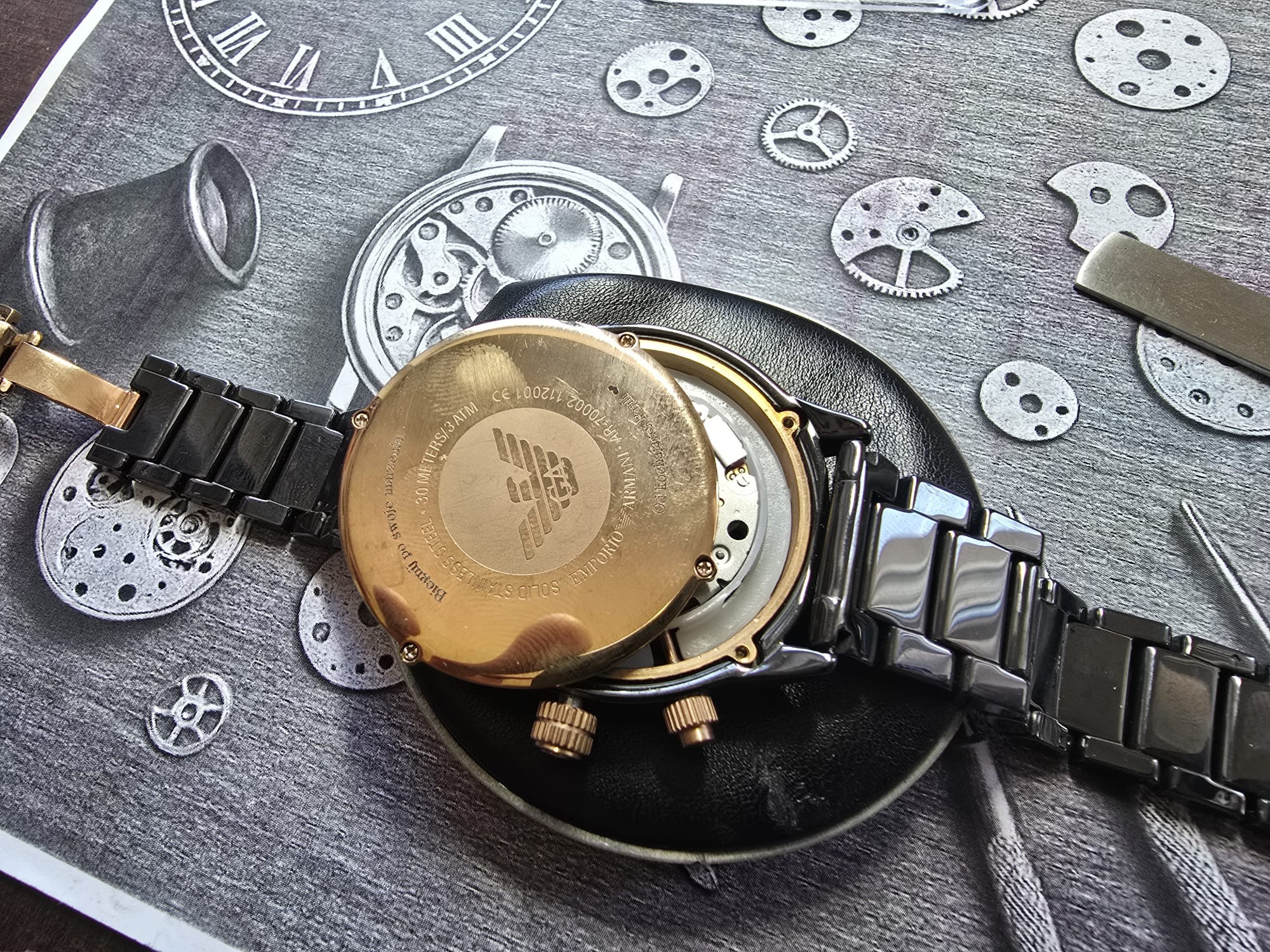 Zegarek Emporio Armani z otwartym deklem na czarnym podłożu z nadrukowanymi elementami zegarków. Widoczne narzędzia zegarmistrzowskie, detale mechanizmu i złote elementy.