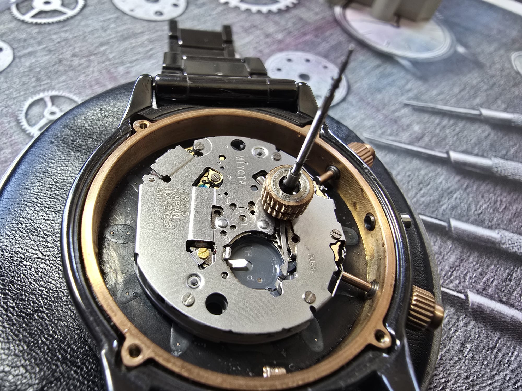 Zegarek kwarcowy bez dekla w trakcie naprawy, widoczny mechanizm i narzędzie zegarmistrzowskie. Tło stanowią elementy zegarków i narzędzia.