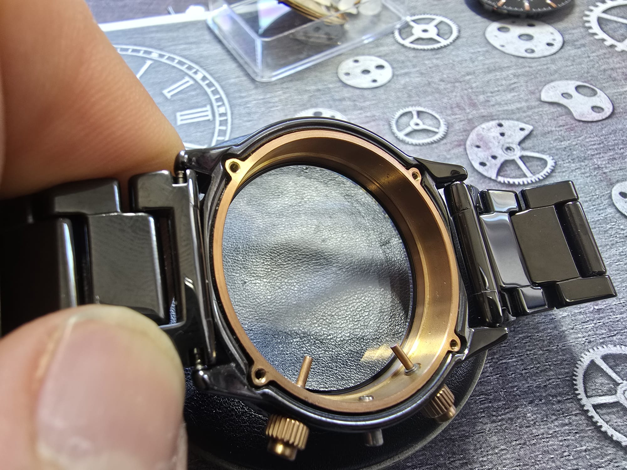 Czarna koperta zegarka bez mechanizmu, trzymana w palcach, na tle z grafiką zegarowych trybików. Widoczne detale: złote pokrętło i mocowania paska.