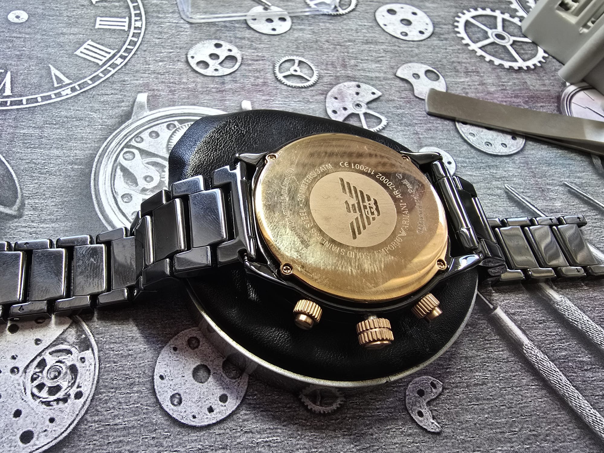 Zegarek Emporio Armani z widocznym deklem i elementami do naprawy na tle z motywem zegarmistrzowskim. Detaliczne ujęcie złotego dekla i narzędzi.