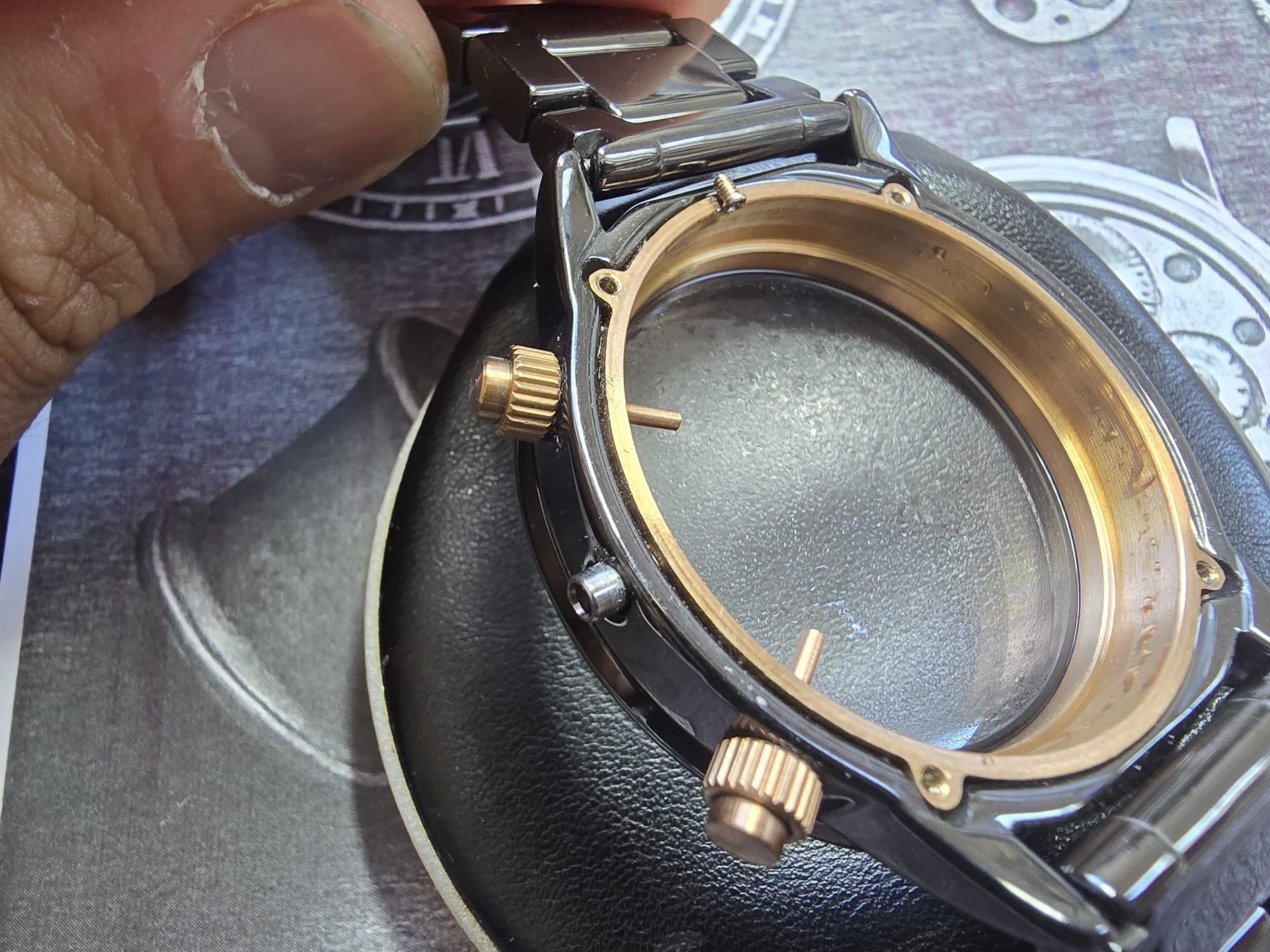 Czarny zegarek bez tarczy, z widocznym mechanizmem, w trakcie naprawy. Palec w lewym górnym rogu. Detal zegarmistrzowski.