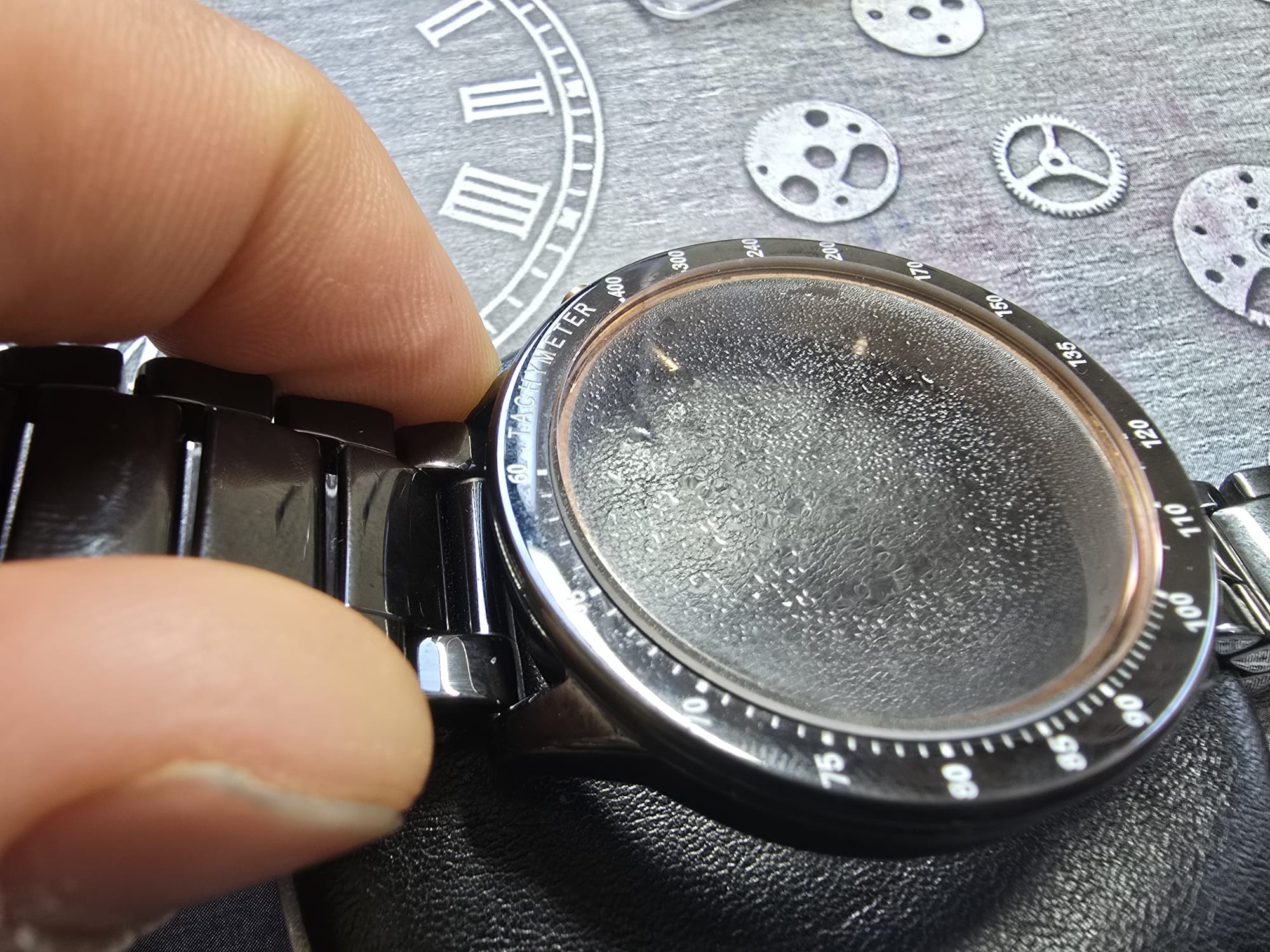 Czarny zegarek z tachymetrem, zamglona tarcza, fragment palca wskazującego, dekoracyjne tło z elementami zegarków. Detal naprawy zegarka.