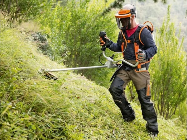 Mężczyzna w ochronnym hełmie i stroju roboczym, używa profesjonalnej kosy spalinowej na nierównym, zarośniętym zboczu wzgórza. Kosa z długim wysięgnikiem.