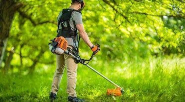 Osoba w kasku i z osprzętem tnie trawę pod drzewami za pomocą podkaszarki spalinowej. Widoczne zielone otoczenie, trawa, drzewa i sprzęt ogrodniczy.