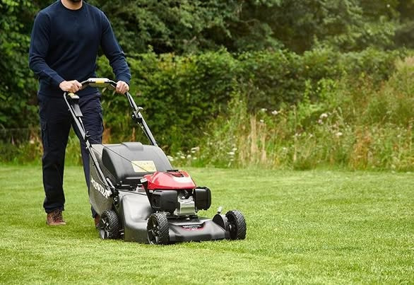 Mężczyzna kosi trawę kosiarką Honda na tle zielonego ogrodu. Krzewy i drzewa w tle. Skoszona trawa równo przycięta.