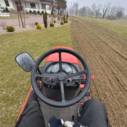 Magiczny Trawnik - Czerwony traktor na zielonym trawniku, widok z perspektywy operatora podczas wykonywania prac ziemnych na posesji z domem w tle.